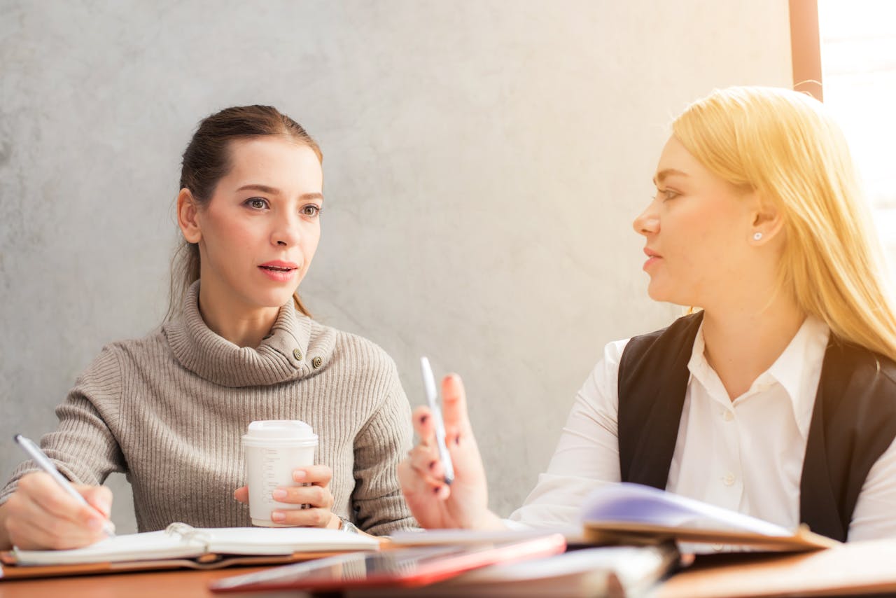 about-us Two women engaging in a professional conversation over coffee and notes.